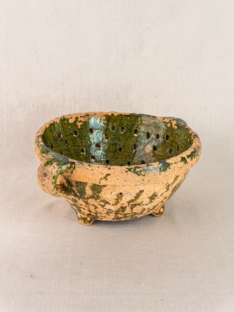 Antique French green glazed terracotta colander with side handles, small feet, and drainage holes on light textured fabric.