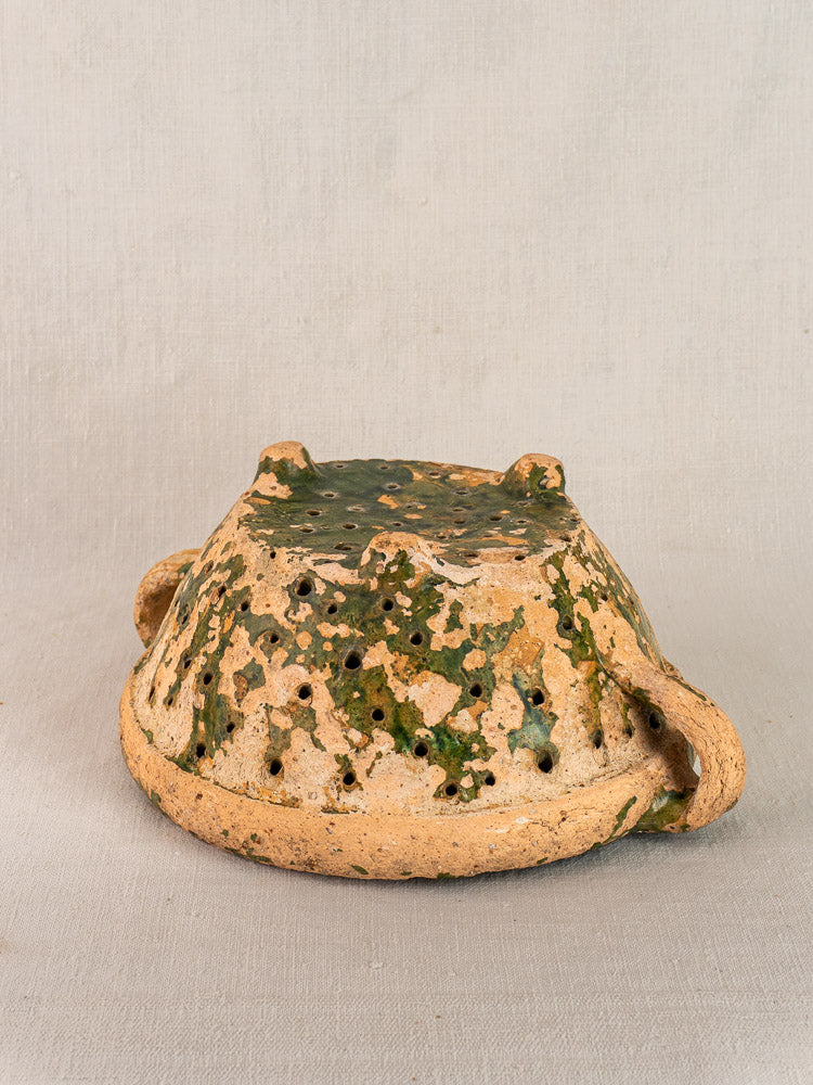 Late 18th-century French terracotta colander with green glaze, earthy brown tones, and curved handle on plain fabric.