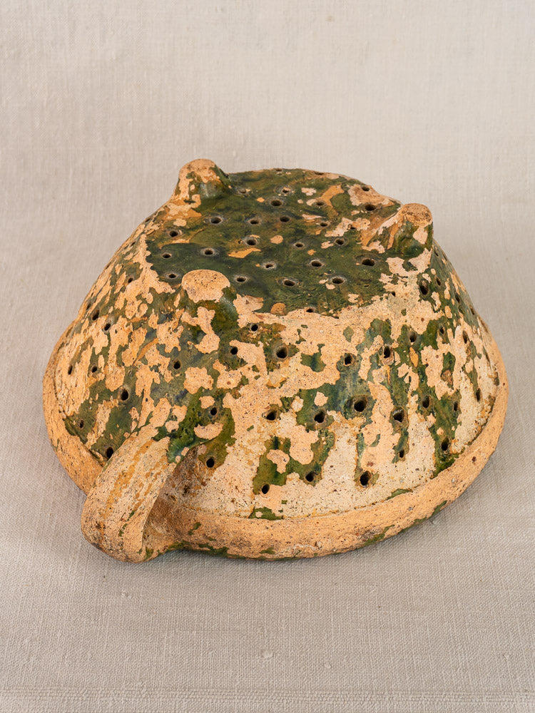 Antique French green-glazed terracotta colander with tan base and perforations on neutral fabric background.