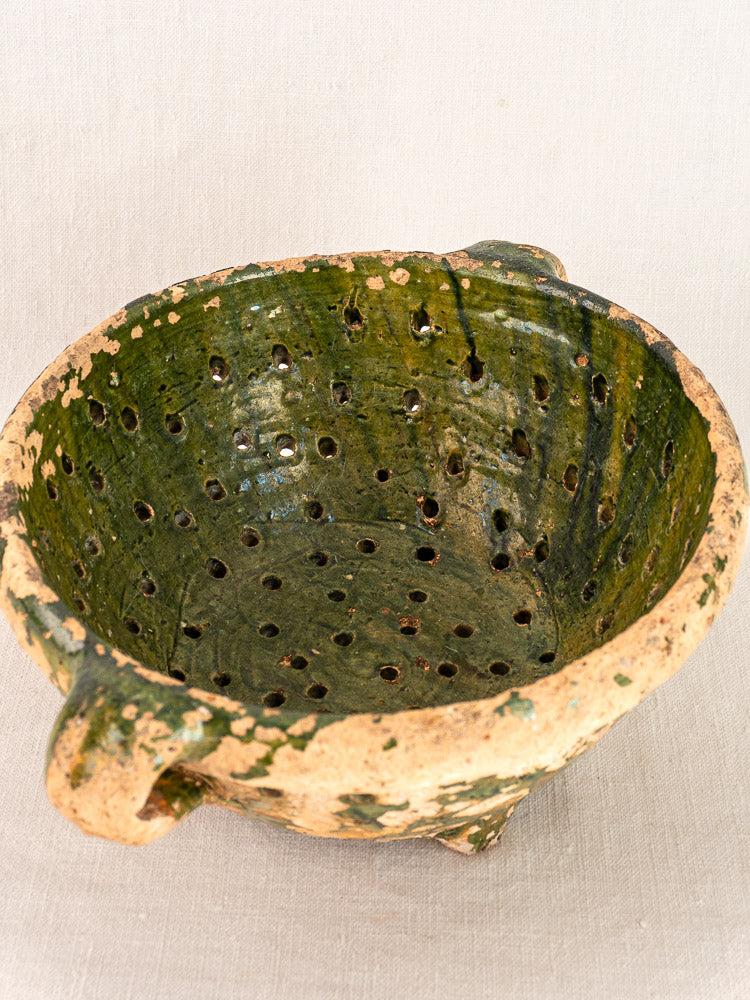 Late 18th century French green glazed terracotta colander with three handles and drainage holes on light fabric.