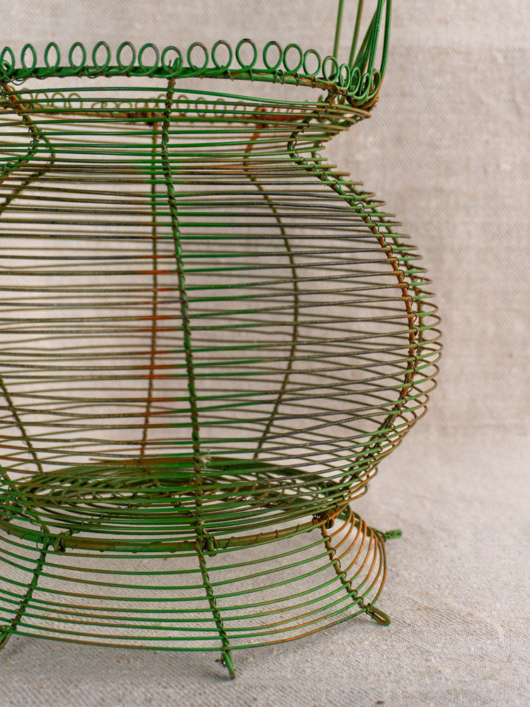 Antique French green wire egg basket with scroll rim and original paint, geometric pattern, on beige fabric background. Mid 20th century.