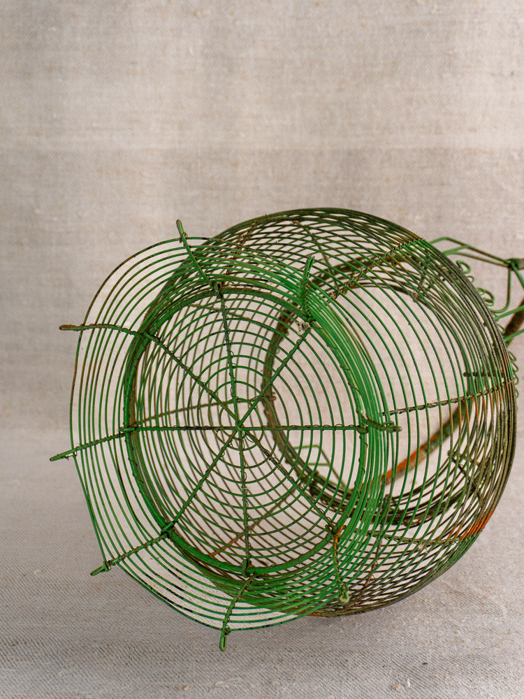 Antique French green wire egg basket with scroll rim and circular base on textured beige fabric.