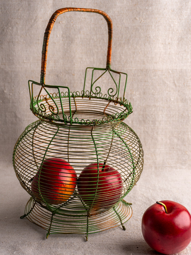 Antique French green wire egg basket with scroll rim, original paint, two red apples inside, one on beige textured surface.