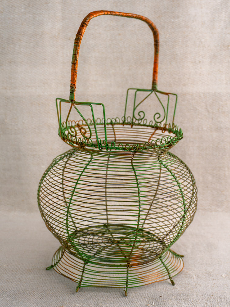 Antique French green wire egg basket with original paint, scroll rim, and orange-wrapped handle on neutral fabric.