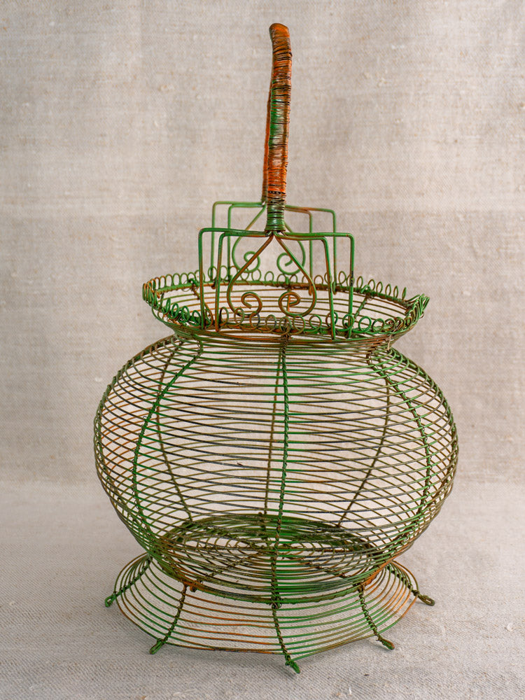 Antique French green wire egg basket with original paint, scroll rim, round body and handle on neutral fabric background.