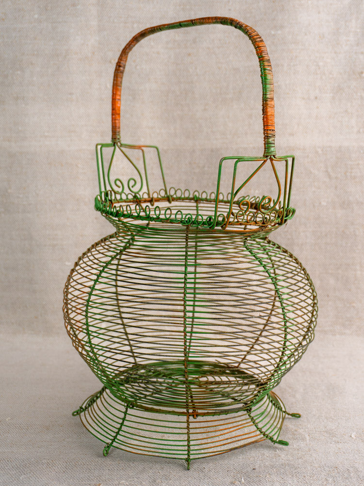 Antique French green wire egg basket with scroll rim, original paint, and upright handle on neutral fabric background.