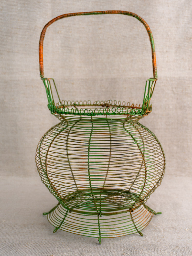 Antique French green wire egg basket, mid-20th century, with scroll rim and handle on neutral fabric background.