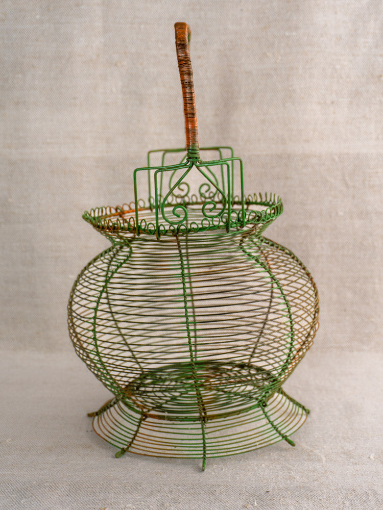 Antique French green wire egg basket with scroll rim and brown-wrapped handle on textured beige fabric.