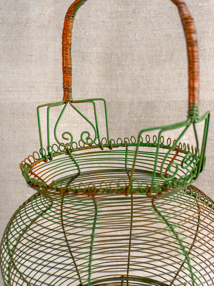 Antique French green wire egg basket with scroll rim and curved handle, mid 20th century, on neutral fabric background.