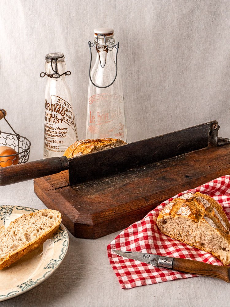 Antique French bread cutter with guillotine blade on wood board, rustic loaf, glass bottles, red napkin.