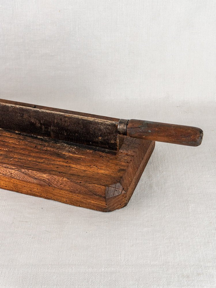 Antique French bread cutter with wooden base and metal blade, displayed on a white textured surface.
