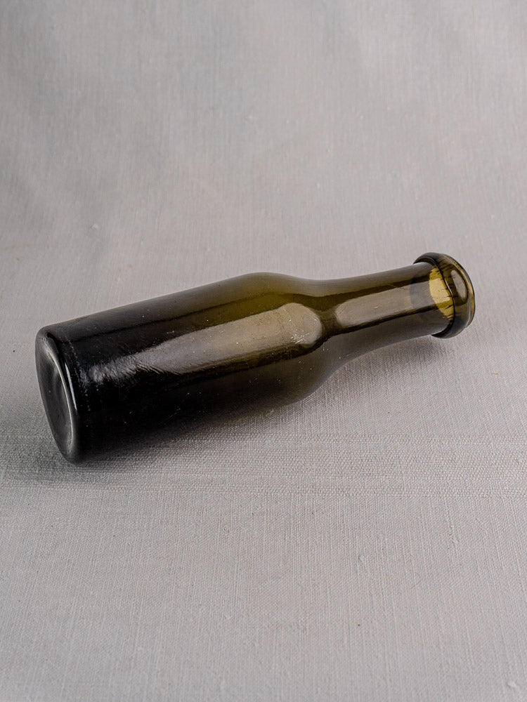 Antique French truffle jar in dark olive green glass resting sideways on light gray textured surface.