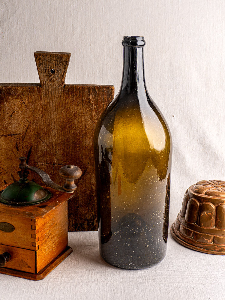Large 19th-century French hand-blown wine bottle in dark olive glass with tall neck, on light textured surface.