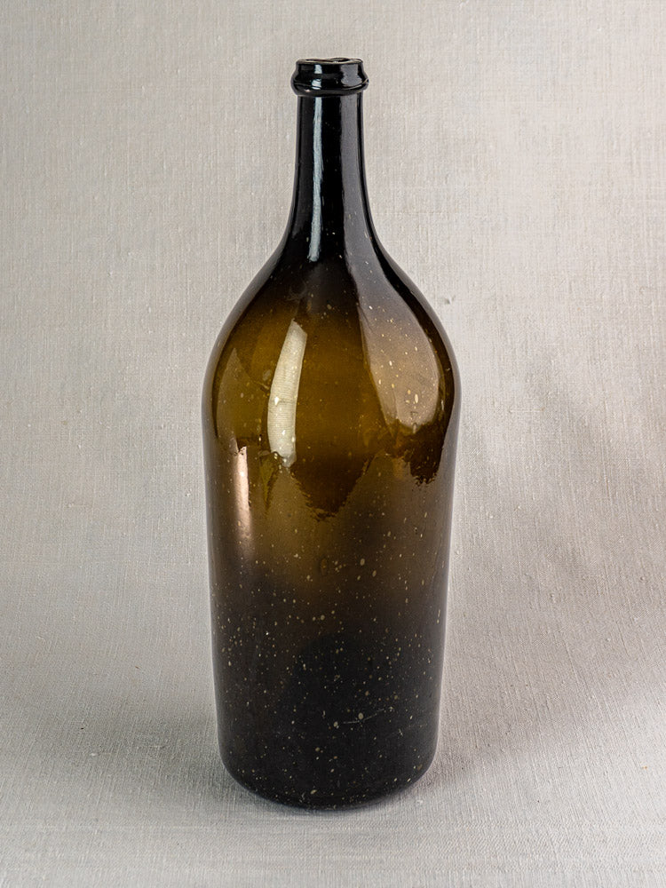 Large 19th-century French hand-blown wine bottle in dark olive glass with tall neck, on light textured surface.