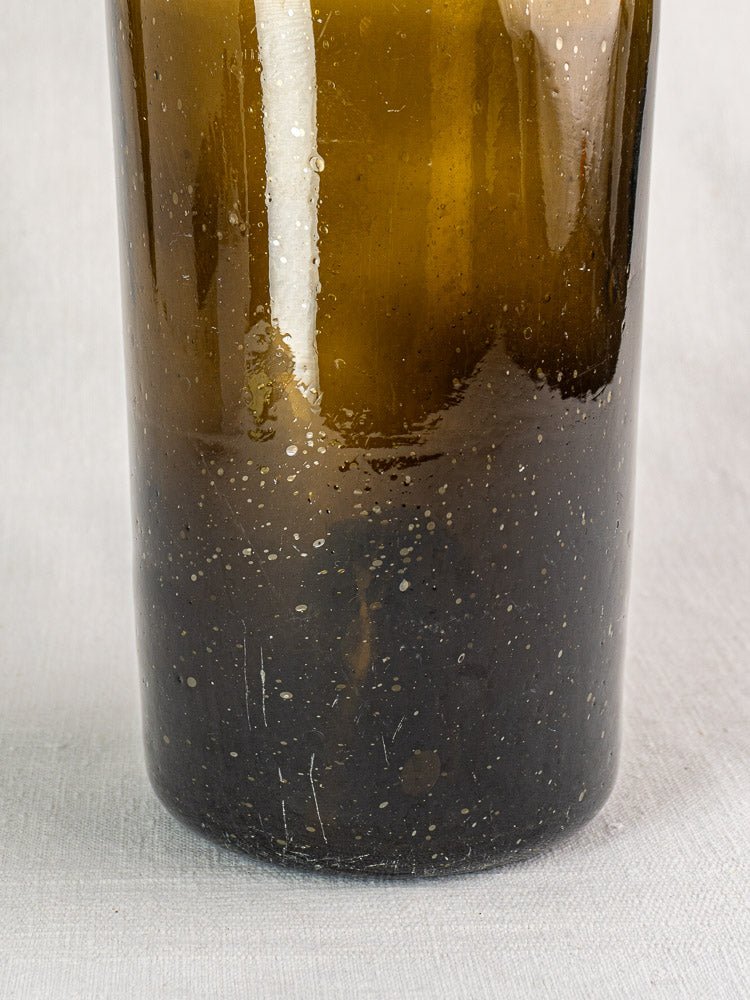 Bottom half of antique French hand blown dark olive glass wine bottle with bubbles and imperfections, on gray fabric.