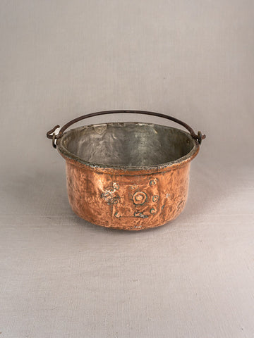 Antique French copper cauldron with repairs, rounded form, weathered patina, and handle on light gray background.