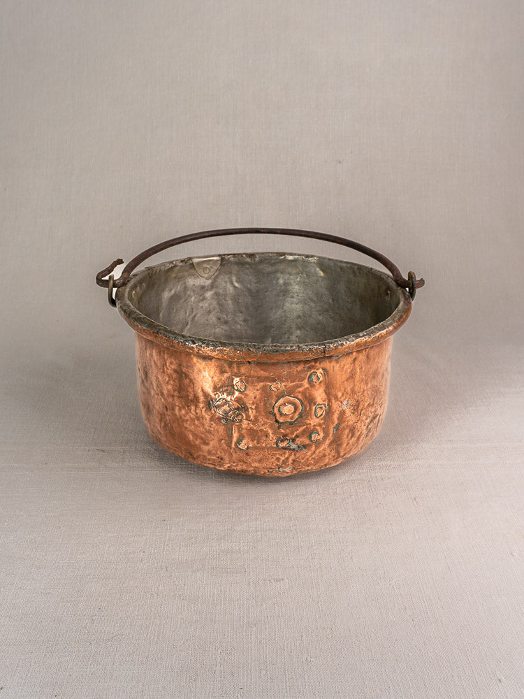 Antique French copper cauldron with repairs, rounded form, weathered patina, and handle on light gray background.