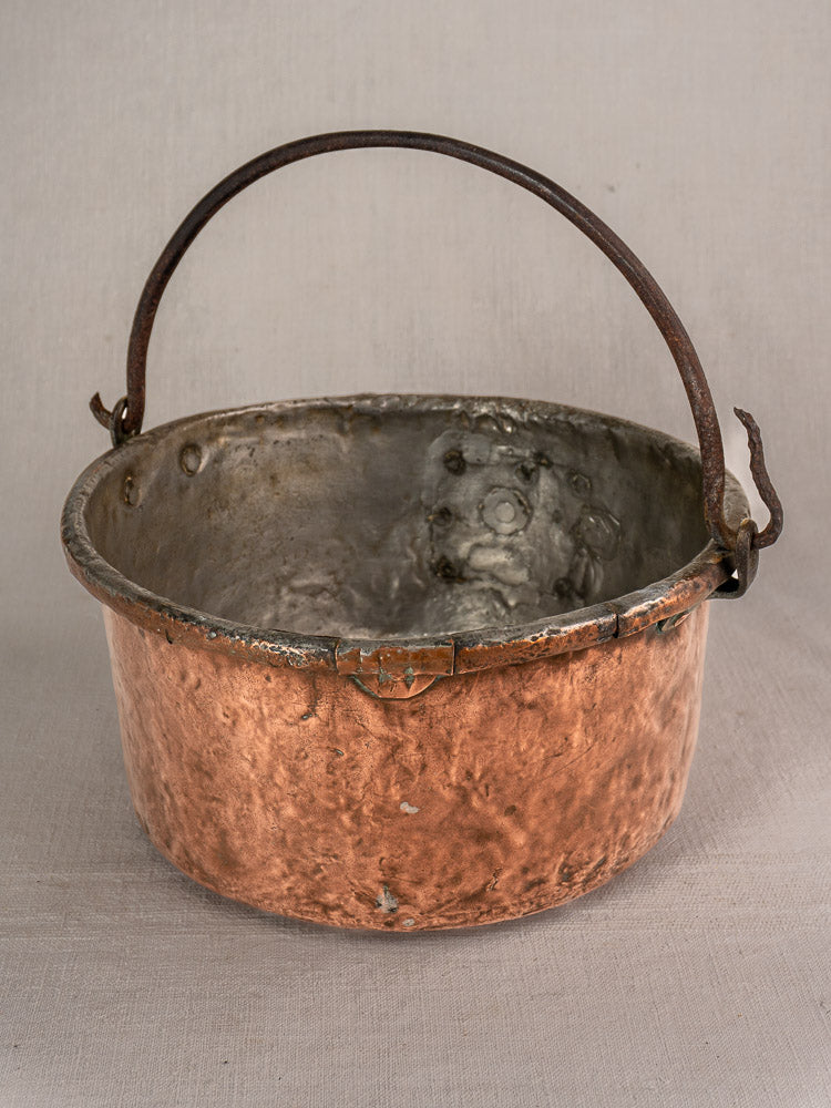 Antique French copper cauldron with round weathered surface, iron handles, and repairs on a neutral fabric background.