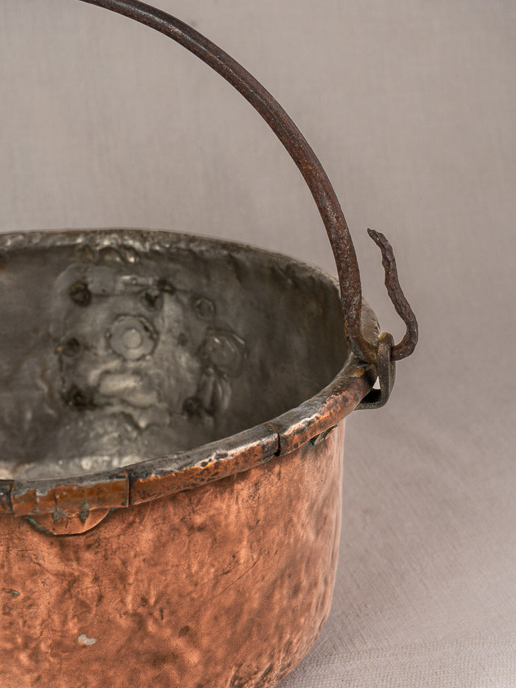 Antique French copper cauldron with hammered surface, iron handle, and repaired interior on neutral fabric background.