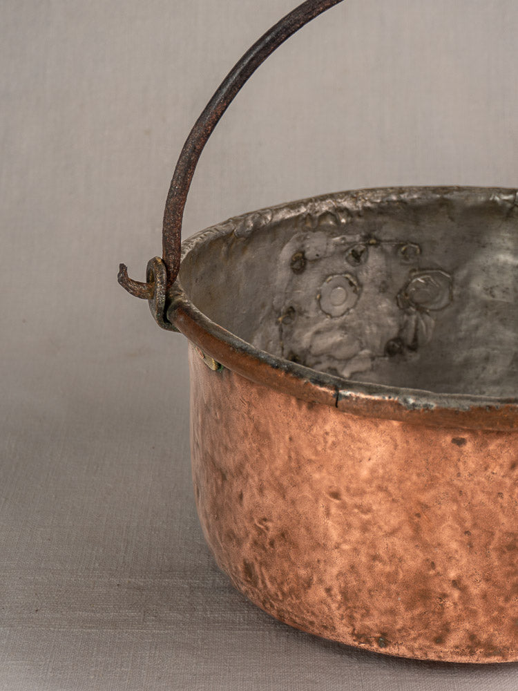Antique French copper cauldron with metal handle, unique repairs, and textured patina on a neutral fabric background.