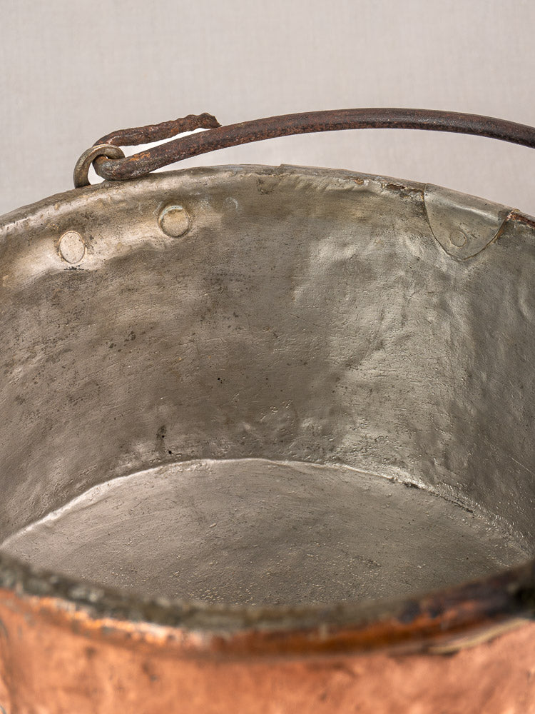 Antique French copper cauldron interior with curved handle, visible rivets, repairs, and aged farmhouse texture.