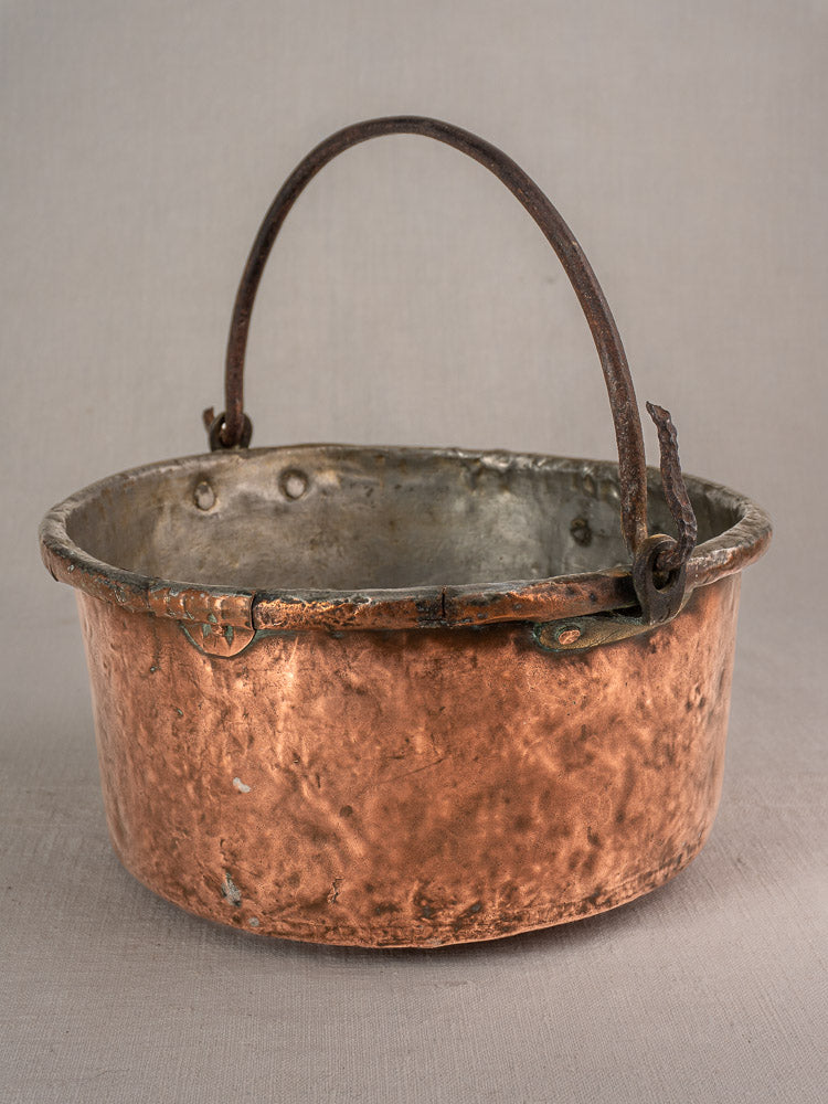 Antique French copper cauldron, 17th–18th century, round with unique repairs and arched rusted handle on gray background.