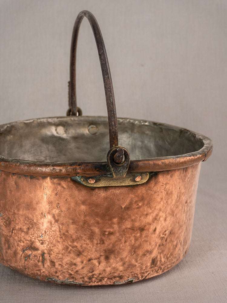 Antique French copper cauldron with metal handle and unique repairs, weathered patina on a neutral surface.