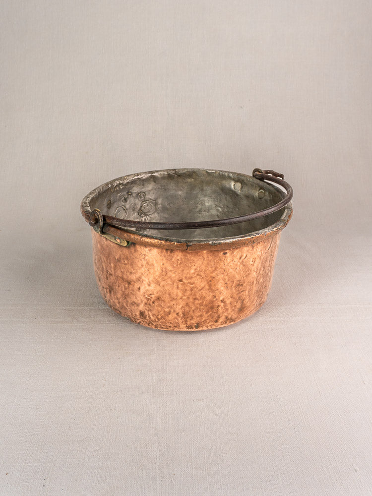 Antique French copper cauldron with round hammered body, metal handle, unique repairs, and aged patina on a light background.