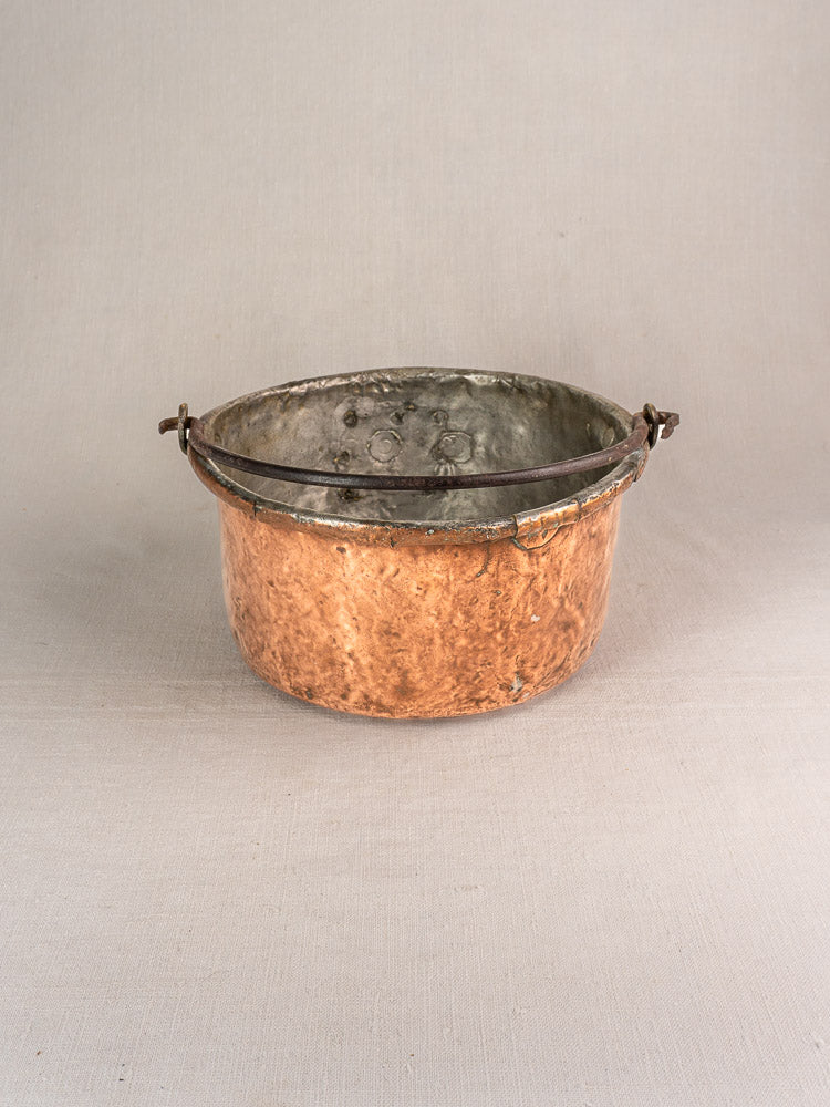 Antique French copper cauldron with dark metal handle and visible repairs displayed on light linen fabric.
