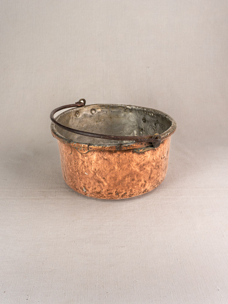 Antique French copper cauldron with metal handle and visible repairs on neutral fabric, highlighting aged patina and texture.