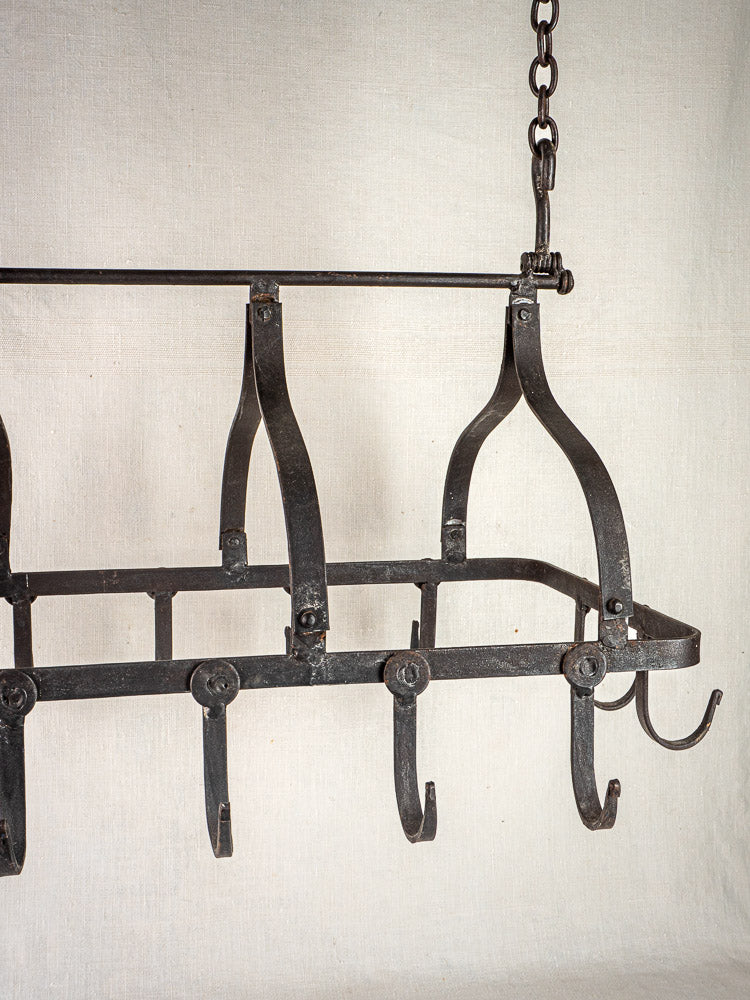 Antique French wrought iron hanging pot rack with curved hooks, suspended by chain, on beige fabric backdrop.