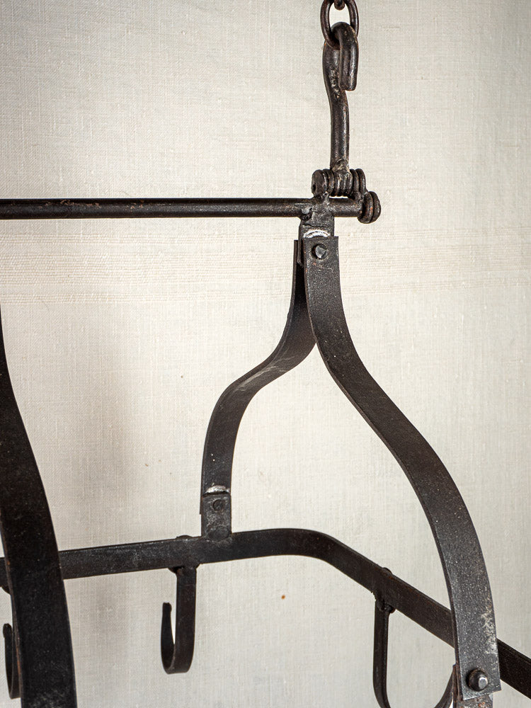 Antique French wrought iron hanging pot rack with S-shaped hooks and curved arms on beige fabric.