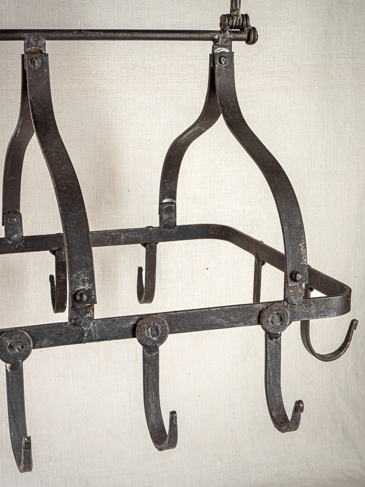 Antique French wrought iron hanging pot rack with curved hooks, set against beige fabric.