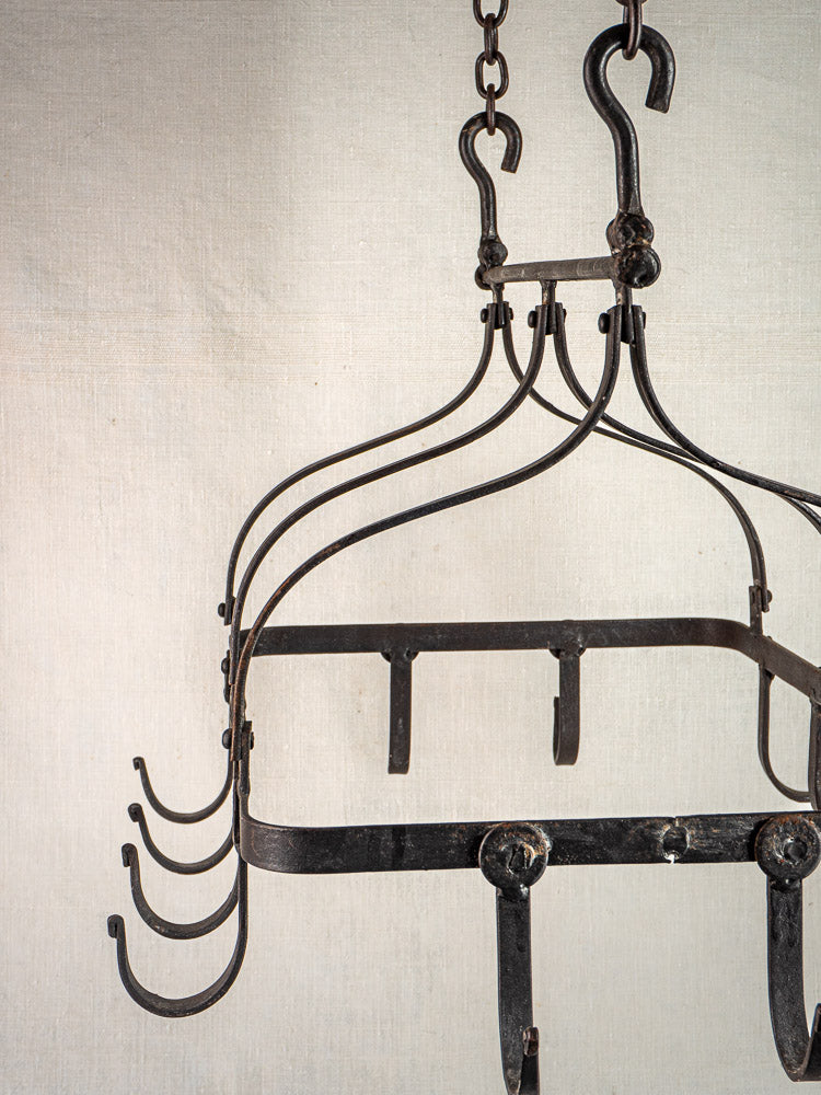 Antique French wrought iron hanging pot rack with multiple hooks and chains on a light fabric background.