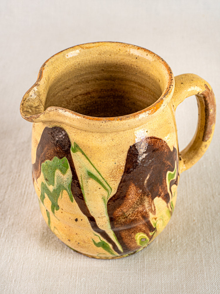 Antique French Jaspé ceramic pitcher with yellowish glaze, green and brown marbled patterns, on neutral surface.