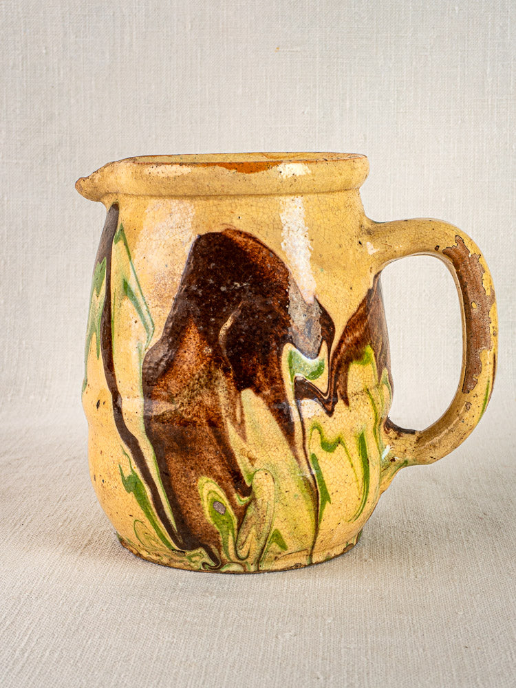 Antique French Jaspé ceramic pitcher with yellowish glaze, green and brown marbled patterns, on neutral surface.