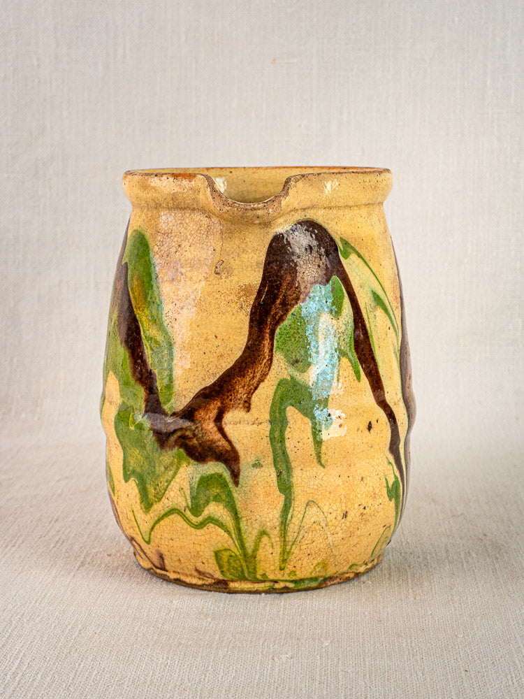 Antique French Jaspé pitcher with tan ceramic body, marbled green and brown glaze, on light fabric background.