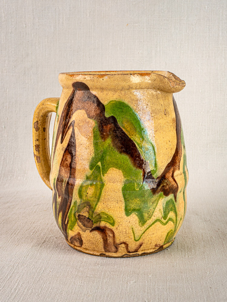 French jaspé ceramic pitcher with handle, yellow base and green-brown marbled glaze on fabric background.