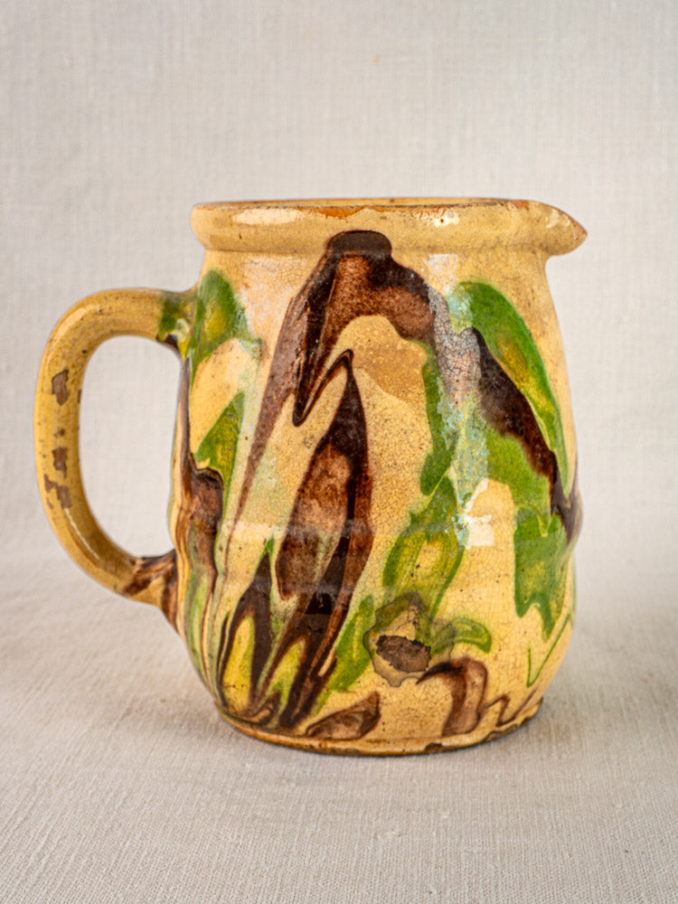 Antique French Jaspé pitcher with marbled green and brown glaze on yellow base, shown on fabric.