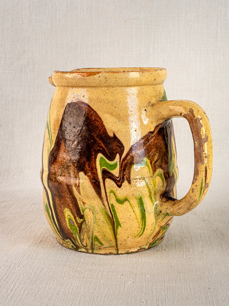 Antique French jaspé ceramic pitcher with marbled brown and green glaze, beige base, on light fabric background.