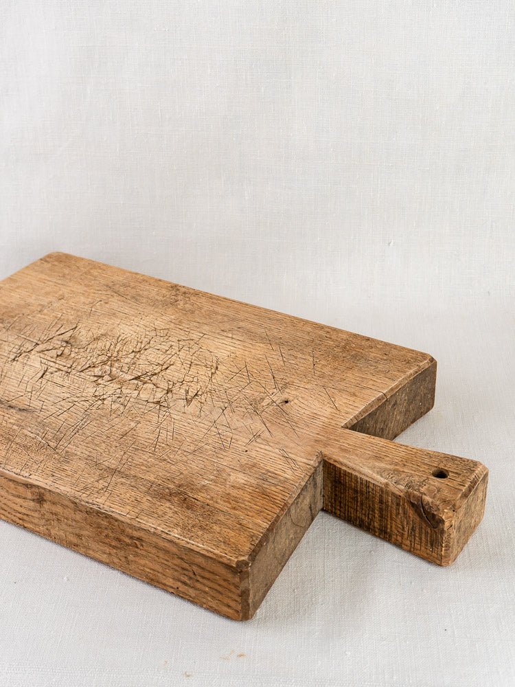 Late 19th-century French oak chopping board with short handle and patina, on light fabric background.