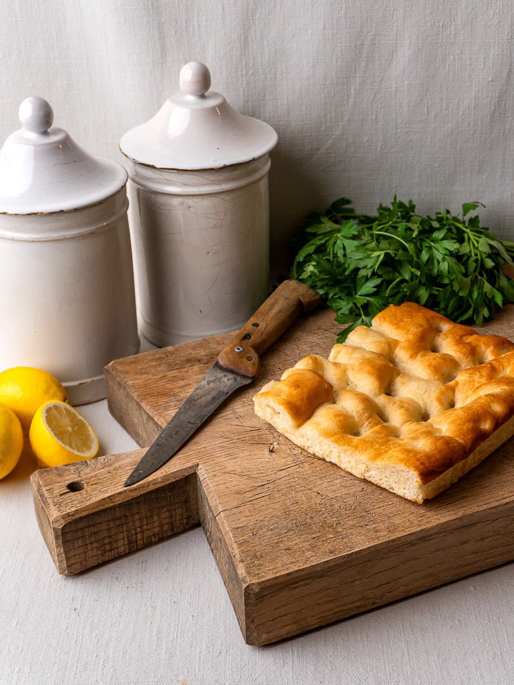 Antique French oak chopping board with focaccia, knife, parsley, lemons, and white ceramic jars.
