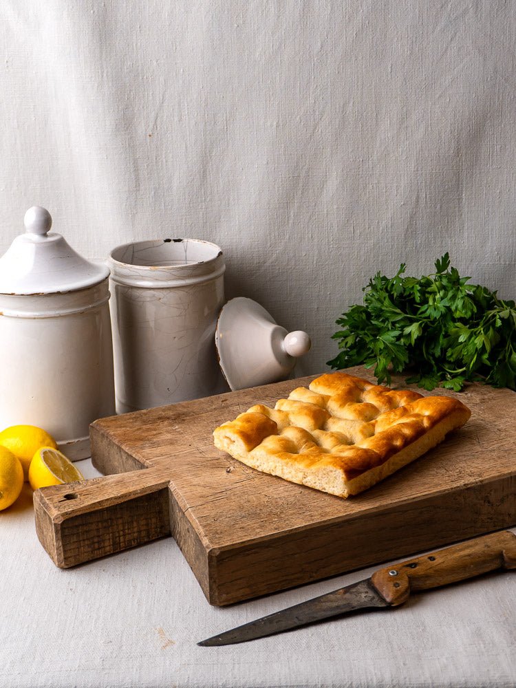 Antique French oak chopping board with focaccia, knife, parsley, lemons, and two white ceramic canisters.