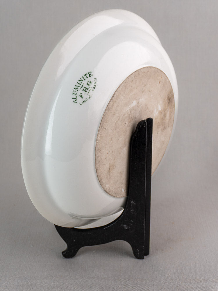 Antique French Aluminite bowl by Frugier Limoges, back view with green mark on black stand, light gray background.