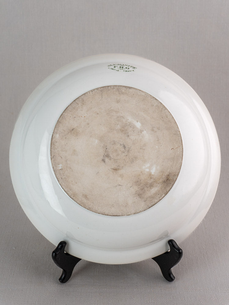 Antique French aluminite bowl by Frugier Limoges upside down on black stand, unglazed base, green mark near rim, gray backdrop.