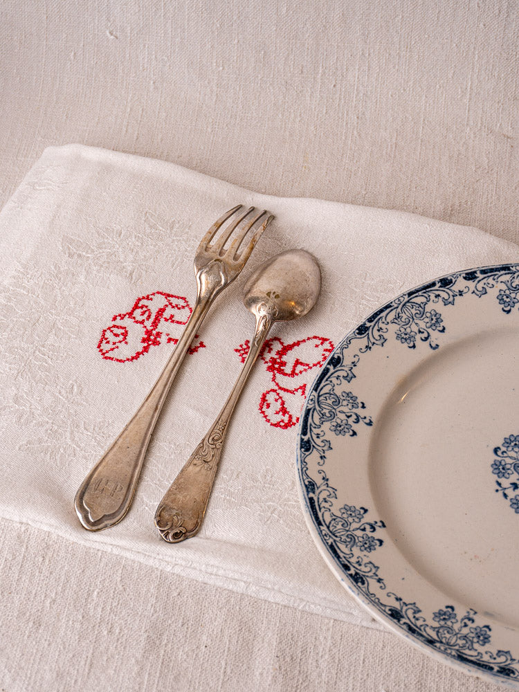 Antique French damask linen napkin with red "P.G." monogram, under vintage flatware and blue floral plate.
