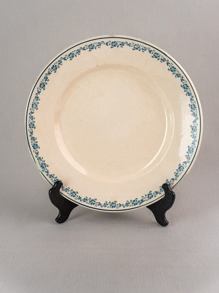 Antique French faience plate, Longwy Capucia pattern, cream base with teal floral border on black stand, gray background.