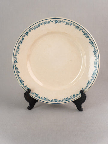 Antique French faience plate, Longwy Capucia pattern, cream base with teal floral border on black stand, gray background.