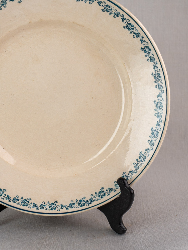 Antique French faience plate in the Longwy Capucia pattern with teal floral border on black stand, gray background.