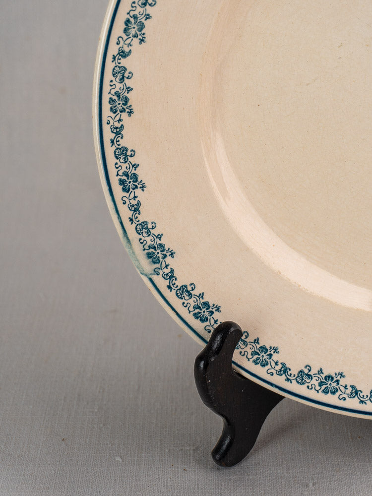 Antique French faience Longwy Capucia plate with teal floral border on dark wood stand, gray backdrop.